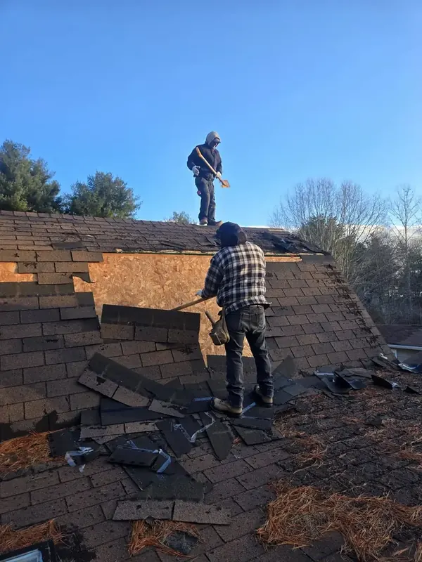 Two workers tearing off old shingles on a steep roof at sunrise.